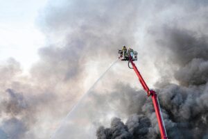 Dramatic scene of firefighters on an aerial ladder battling dense smoke from a fire.